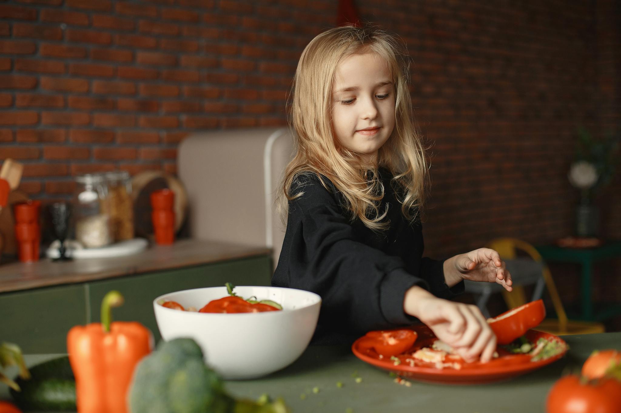 Cheerful little blond child preparing salad while removing pepper seeds and standing near table with fresh vegetables behind brick wall in apartment