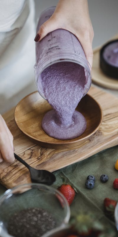 Close-up of a berry smoothie being poured into a wooden bowl, surrounded by fresh ingredients.