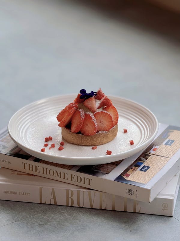 Elegant strawberry tart on a white plate atop stacked books, showcasing chic food styling.