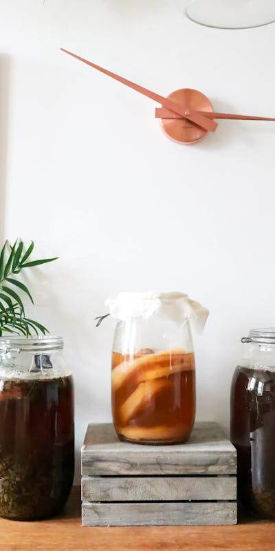 Glass jars with infused liqueurs with ingredients placed on wooden table near green plant near wall with decorations at home