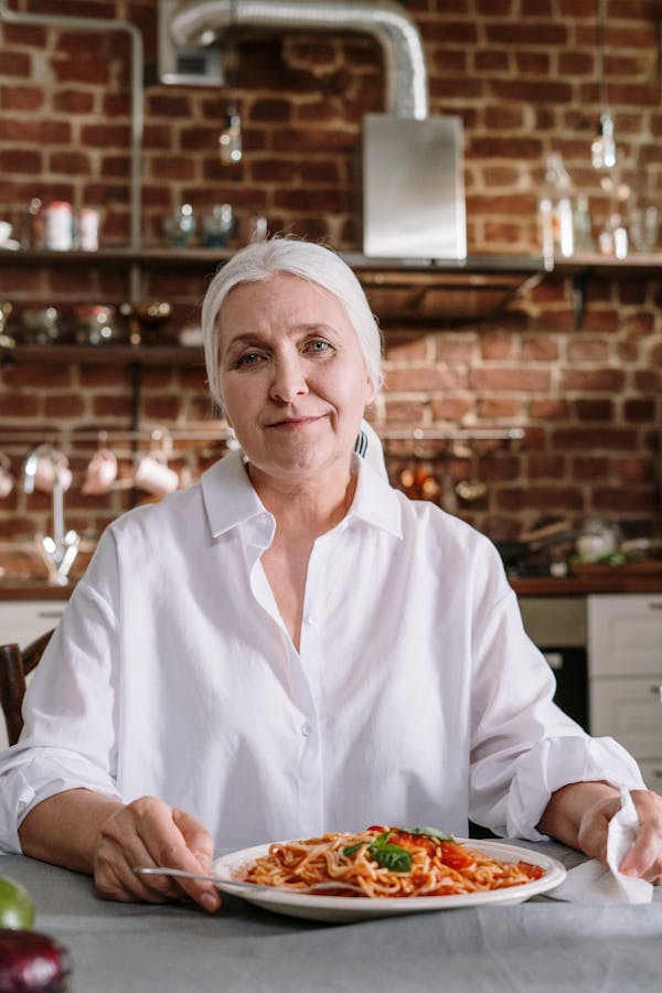Senior woman with grey hair smiling at a table with a plate of spaghetti in a rustic kitchen.